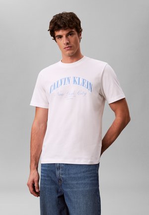 Młody mężczyzna ubrany w białą koszulkę Calvin Klein z niebieskim napisem "New York City", w połączeniu z niebieskimi dżinsami, stojący na jednolitym szarym tle.