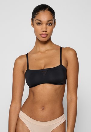 Kobieta o ciemnych plecionych włosach, ubrana w czarny krótki top na cienkich ramiączkach i jasnobeżową bieliznę, stojąca na tle jednolitego tła.