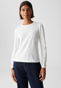 Donna con capelli corti scuri che indossa una maglietta bianca a maniche lunghe e jeans blu scuro, in piedi contro uno sfondo chiaro e uniforme.