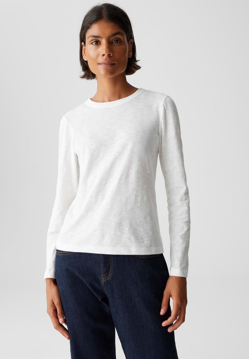 Donna con capelli corti scuri che indossa una maglietta bianca a maniche lunghe e jeans blu scuro, in piedi contro uno sfondo chiaro e uniforme.