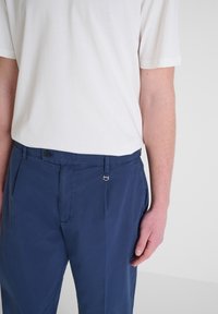 Pantaloni blu navy con una texture liscia, dotati di chiusura a bottone e passanti per cintura. Abbinati a una camicia a maniche corte bianca semplice.