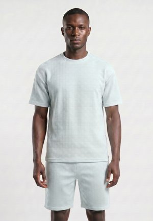 Homme portant une chemise à manches courtes texturée bleu clair et un short assorti, debout devant un fond uni.