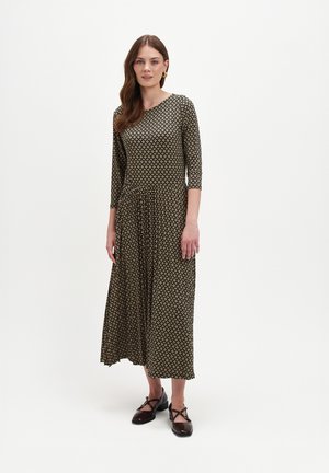 Kobieta stoi w midi sukience z długim rękawem i wzorem, zestawionej z ciemnobrązowymi butami typu Mary Jane, na jednolitym białym tle.