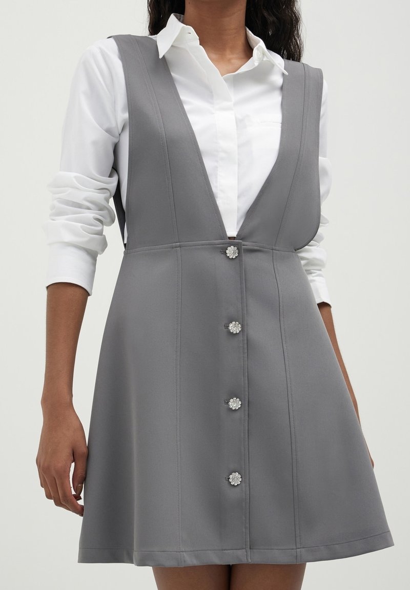 Femme portant une chemise blanche à manches longues sous une robe sans manches grise avec des boutons décoratifs sur le devant.