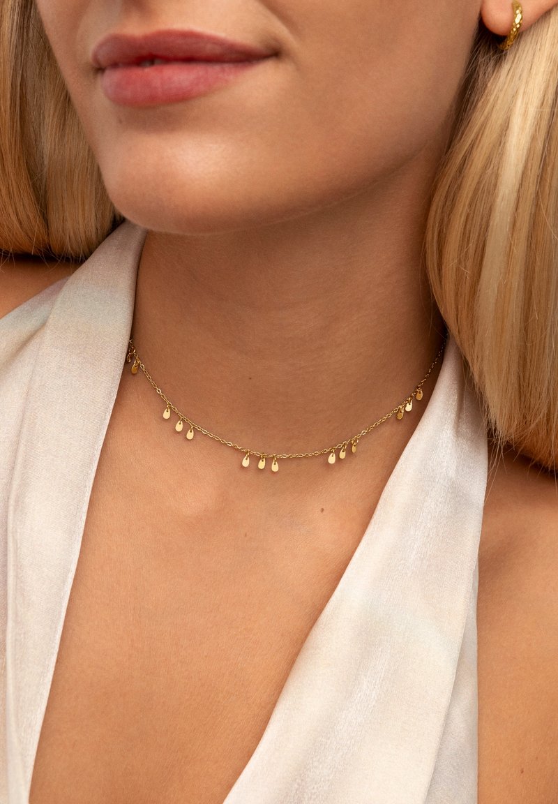 Collana choker in oro con pendenti a forma di goccia su una delicata catena, indossata su un collo scoperto su uno sfondo di tessuto chiaro.