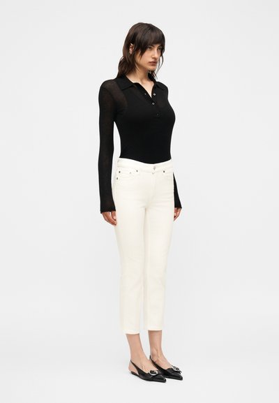 Γυναίκα στέκεται με το πρόσωπο προς τα εμπρός, φοράει μαύρο μακρυμάνικο πόλο, λευκό ψηλόμεσο κροπ παντελόνι και μαύρες μυτερές πλατφόρμες slingback.