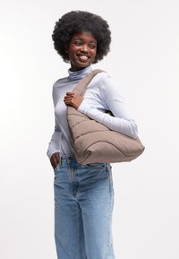Torba puffer w kolorze taupe, o zaokrąglonym kształcie i szerokim pasku. Połączona z jasnoniebieską koszulą z długim rękawem i dżinsami z wysokim stanem.