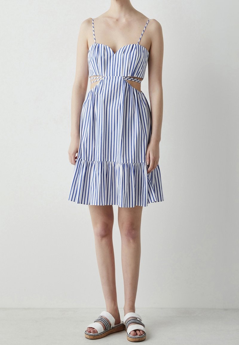 Femme portant une robe sans manches rayée verticalement bleu et blanc avec des découpes à la taille et des sandales blanches à enfiler.