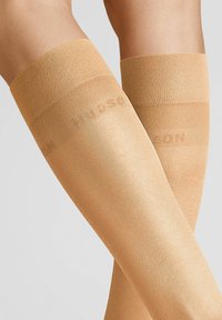 Beige Oberschenkelstrümpfe mit glatter Textur und breitem Gummiband oben. Dezenter Glanz und Branding auf dem Band sichtbar.