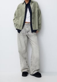 Camisa de pana verde de gran tamaño sobre una camisa azul marino a rayas, combinada con jeans de pierna ancha gris claro y zapatos negros sin cordones.