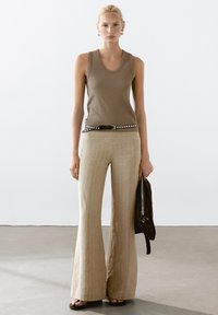 Bruine tanktop, beige gestreepte wijde broek, zwarte gespietste riem, zwarte tas in de hand, platte sandalen, lichte achtergrond, minimalistisch ontwerp.