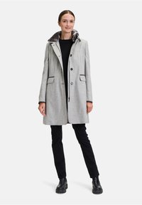 Grau wollener Mantel mit einem Kunstpelzkragen, ausgestattet mit einer Knopfleiste, Seitentaschen und einer taillierten Form. Kombiniert mit schwarzen Hosen und Stiefeln.