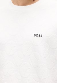 Κοντινό πλάνο ενός λευκού υφασμάτινου πουκαμίσου με μικρό μαύρο λογότυπο "BOSS" κεντημένο στο στήθος κοντά στον λαιμό.