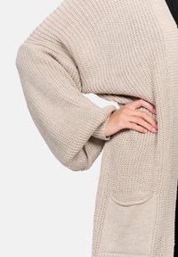 Beige knitted cardigan with wide sleeves, ribbed texture, and a pocket on the front. The fabric appears soft and slightly chunky.