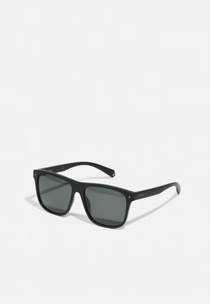 Óculos de sol pretos em formato angular com lentes cinzas escuras. Aros com acabamento mate, hastes largas e uma marcação discreta nas pernas.