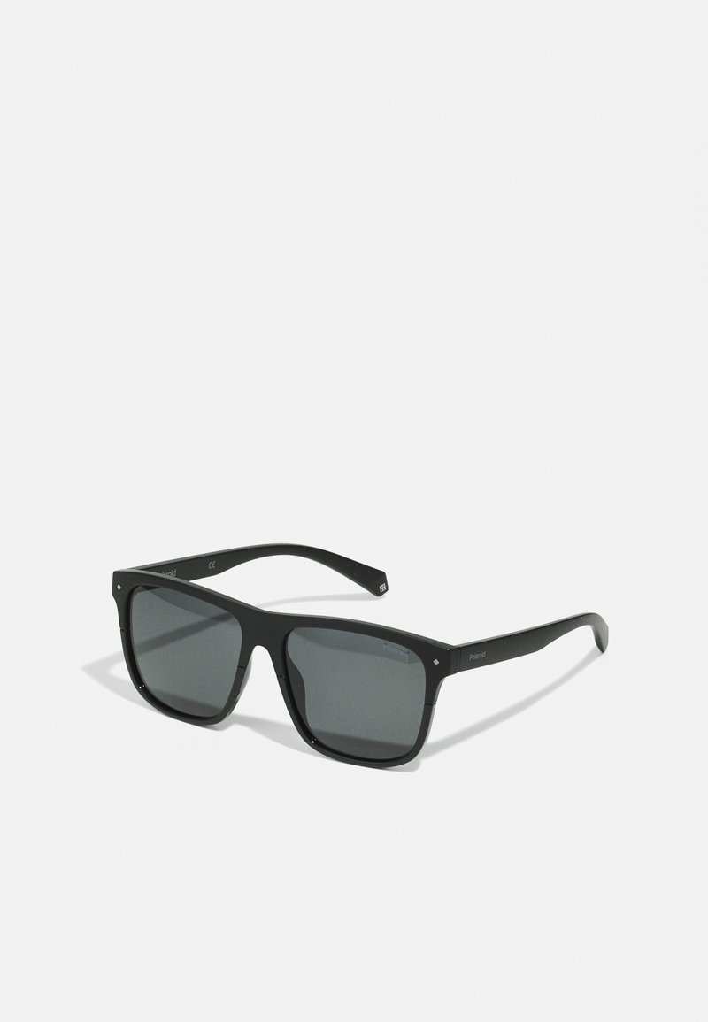 Gafas de sol negras de forma angular con lentes gris oscuro. Montura con acabado mate, patillas anchas y un sutil logotipo en los brazos.