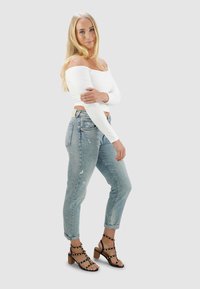 Witte off-shoulder top met lange mouwen, ver distressed lichtblauwe jeans, en zwarte sandaalhakken met gouden studs op de bandjes.