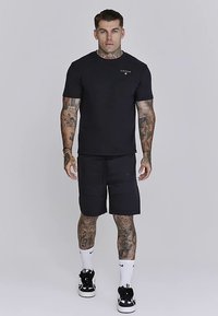 Czarny t-shirt z krótkim rękawem z małym logo na piersi, w połączeniu z czarnymi szortami. Noszony z czarno-białymi tenisówkami i białymi skarpetkami.