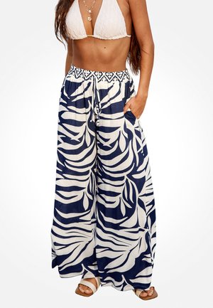 Femme portant un pantalon à jambes larges avec un imprimé de feuilles tropicales bleu marine et blanc, associé à un haut de bikini blanc texturé et des colliers superposés.