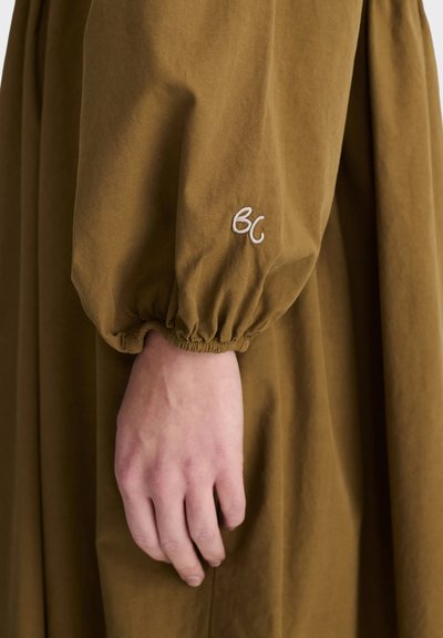 Main avec un teint clair contre une manche en tissu marron avec un poignet élastique et des lettres blanches brodées "BC".