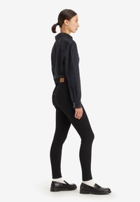 Žena stojí v čiernej denimovej košeli, čiernych slim nohaviciach, bielych ponožkách a čiernych mokasínach, čelí doprava na bielom pozadí.