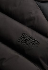 Tejido de chaqueta aislante negra con el logo "SUPERDRY" en relieve. Textura suave y construcción entallada con detalles de costura.