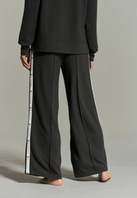 Zwarte wijde broek met ribbelstructuur, met witte zijstrepen en logo-accenten. Gepaard met een bijpassend zwart topje. Blootsvoets.