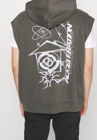 Sudadera sin mangas de color gris oscuro con un gráfico abstracto blanco de una casa, un objetivo, alambre de pua y el texto "ACCHFERRATON" en la parte de atrás.