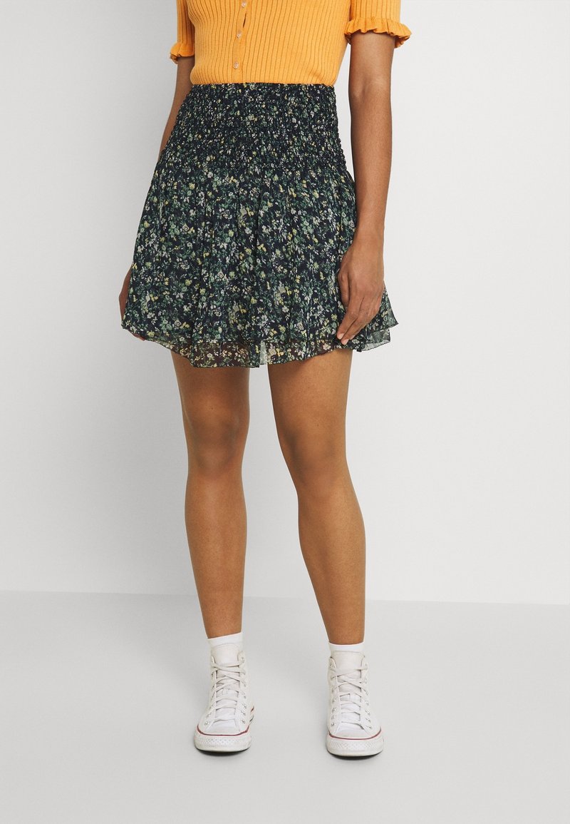 Femme portant une mini-jupe noire à motif floral, un haut à manches courtes côtelé orange, des chaussettes blanches et des baskets montantes blanches.