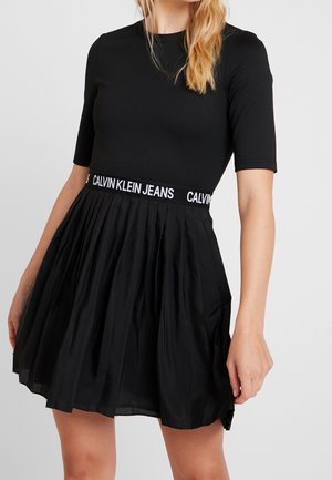 Vrouw draagt een zwarte jurk met korte mouwen, een geplooide rok en een tailleband met het Calvin Klein Jeans-logo, staand tegen een effen achtergrond.