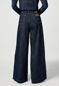 Henkilö, joka wear tummansinisiä leveälahkeisia farkkuja ja niihin sopivaa farkkutakkia, seisoo kädet lanteilla tasaisella taustalla.