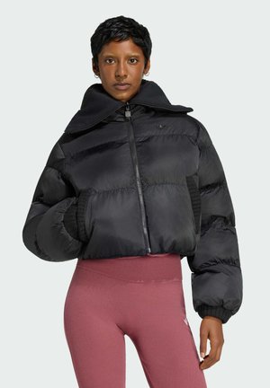 Vrouw met kort haar die een zwart pufferjack met hoge kraag en roze hoge taille leggings draagt tegen een effen achtergrond.