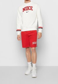 Červené sportovní šortky s logem "NIKE", doplněné bílou mikinou se červeným logem a pruhovanými manžetami. Doplňuje bílé vysoké tenisky.