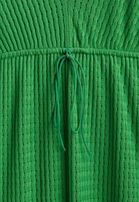 Un tissu texturé vert présente des rayures ondulées en relief avec une fermeture à cordon à la taille, créant un effet froncé.