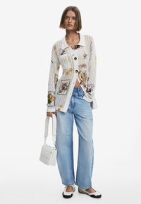 Balta megzta kardiganas su spalvingais gėlių raštais ir rudais mygtukais, derinamas su šviesiai mėlynomis plačiomis kelnėmis ir juodais bei baltais slip-on batais.