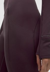 Temno vijolična športna majica z dolgimi rokavi, gladko teksturo in ravnimi šivi, poudarjenega kroja s skozenjimi odprtinami za palce pri manšeti.