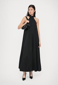 Femeie îmbrăcată într-o rochie lungă neagră fără mâneci, cu un top de tip halter, ținând o mică geantă neagră, purtând pantofi cu toc ascuțit negri, stând pe un fundal alb.