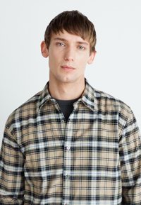 Light brown plaid shirt featuring dark and light blue accents, button-down collar, and a relaxed fit. Smooth cotton texture visible.