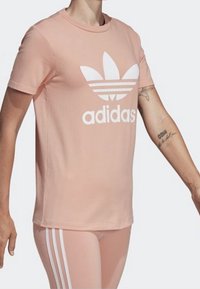 Person som bär en ljusrosa Adidas T-shirt med stort vitt logotyp och matchande leggings med vita ränder på sidorna, arm-tatuering synlig.