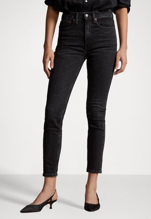 Frau trägt schwarze Skinny-Jeans mit hohem Bund und schwarze Slingback-Schuhe mit niedrigem Absatz, steht vor einem schlichten Hintergrund.