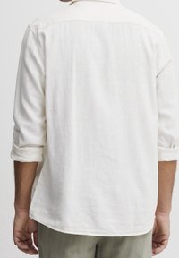 Chemise en lin blanche à manches retroussées, avec une coupe décontractée et un ourlet incurvé. Texture lisse, design simple et sans motifs visibles.