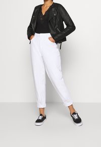 Giacca da moto in pelle nera su un body in pizzo nero, abbinata a pantaloni bianchi arrotolati e sneakers nere con suole bianche.