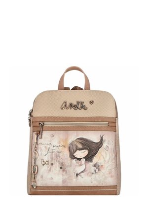 Sac à dos beige en matériau texturé, avec une poche avant ornée d'un design illustré et de breloques en métal. Comprend deux poignées de transport supérieures.