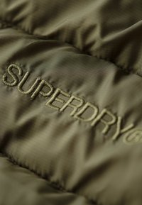 Chaqueta acolchada de color verde oliva con textura acolchada. El logo "SUPERDRY" en bordado tonal añade detalle. Acabado suave y brillante.