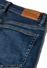 Tamsiai mėlynos džinsinės kelnės su tekstūruotu paviršiumi, turinčios rudos odos ženklelį su "HEYANNO" logotipu ir kontrastingais siūlų detalėmis.