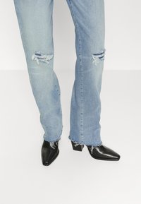 Jeans de mezclilla desgastados en un azul claro con bajos deshilachados, combinados con botas de tobillo negras de punta afilada con tacón ancho.