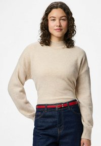 Jeune femme aux cheveux bouclés portant un pull beige, un jean foncé et une ceinture rouge, debout avec une main sur la hanche.