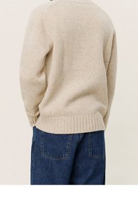 Beżowy sweter robiony na drutach z ribbowym dołem, o chropowatej fakturze. W połączeniu z niebieskimi dżinsami, pokazany z tyłu.