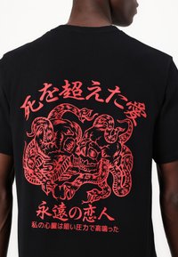Čierne bavlnené tričko s veľkým červeným grafickým motívom s lebkou a hadmi, doplnené japonským textom. Okrúhly výstrih a krátke rukávy.