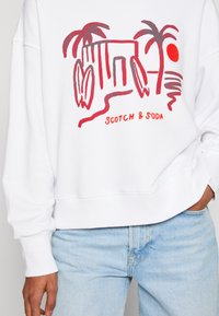 White sweatshirt featuring a red graphic design with a building, palm trees, and sun. Ribbed cuffs and waistband, relaxed fit.